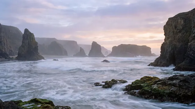 Rocky sea stacks and mossy boulders under a pastel sunset sky, with waves blurred into silky white mist.