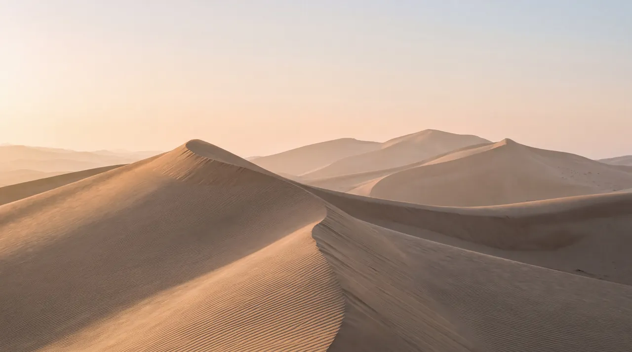 Rippled sand dunes with a sharp crest in the foreground, fading into hazy peach-colored dunes and sky.