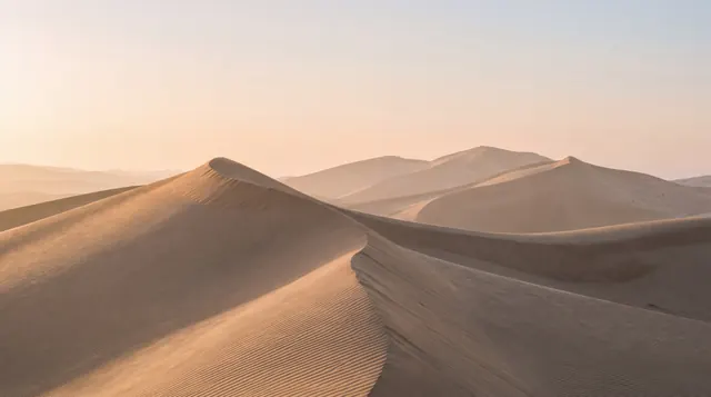 Rippled sand dunes with a sharp crest in the foreground, fading into hazy peach-colored dunes and sky.